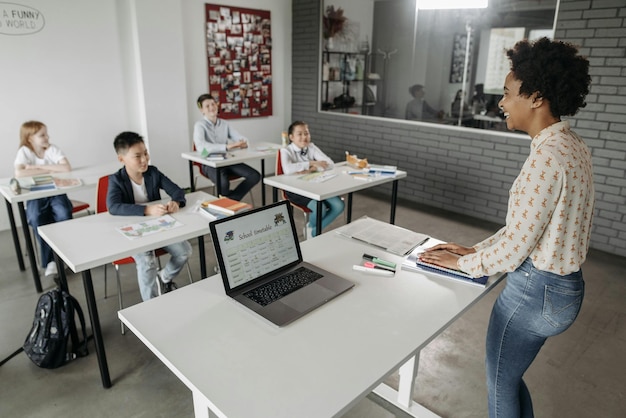A teacher using a laptop at their desk, with books and other teaching materials neatly organized around them. A student can be seen blurred in the background, indicating a classroom environment.